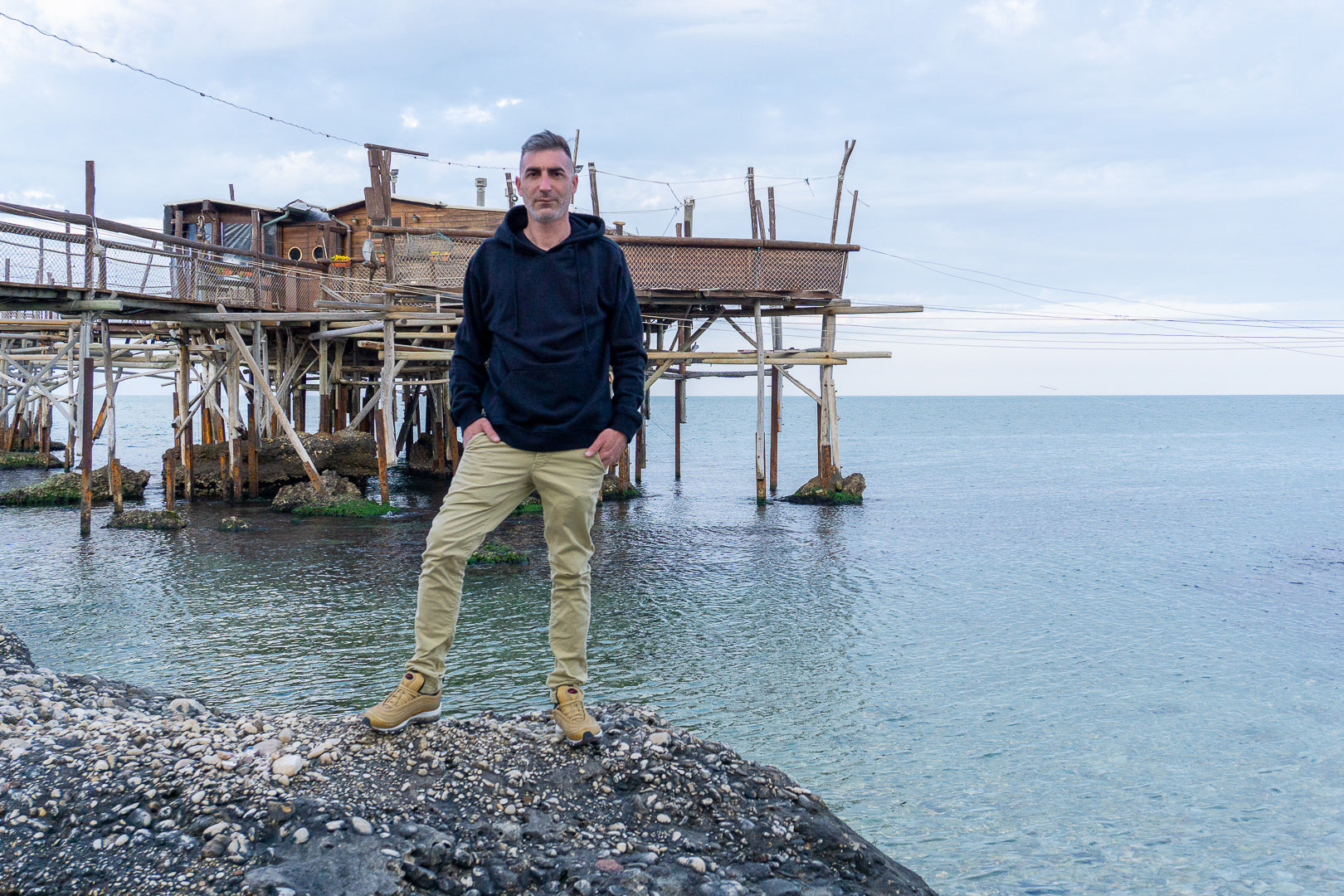 Uomo indossa felpa nera ecologica in bambù davanti a trabocco sul mare - EcoZone abbigliamento sostenibile